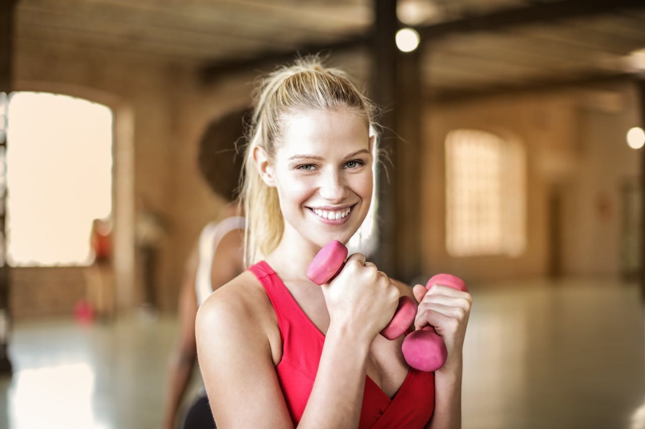 A smiling woman holding pink dumbbells in a bright gym setting, portraying fitness and joy.
